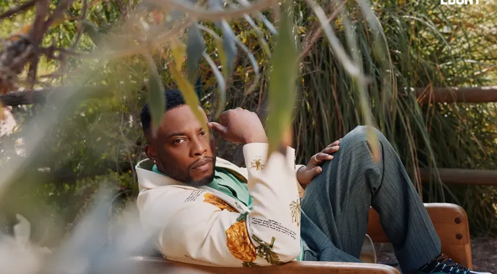 Man leaning on a wooden bench, wearing a light-colored hoodie. Surrounded by foliage, sunny outdoor setting.