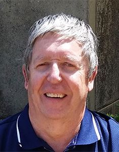 Man with graying hair smiling, wearing a dark blue polo shirt outdoors.