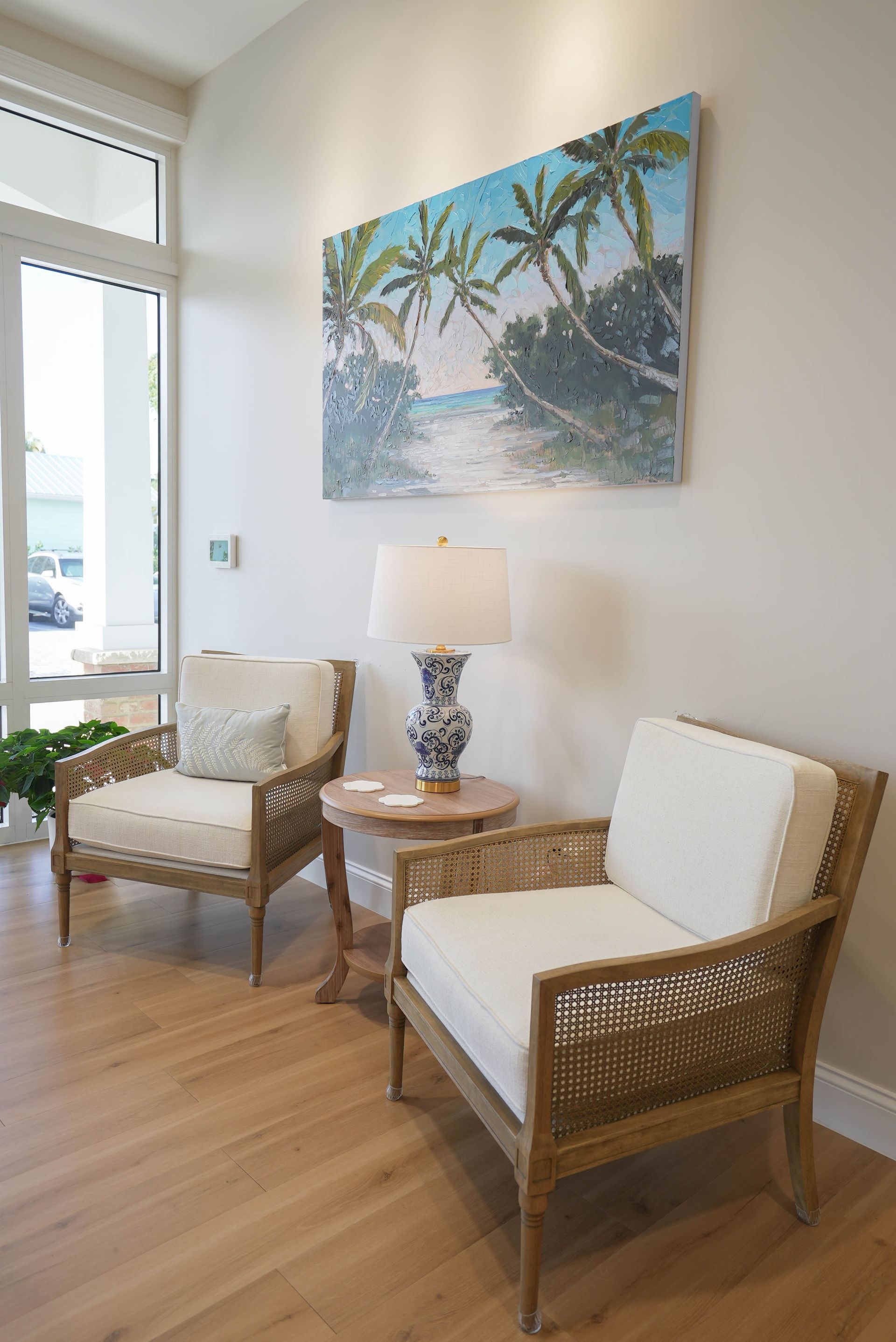 Two wicker chairs with white cushions face each other near a small table and a painting of palm trees.