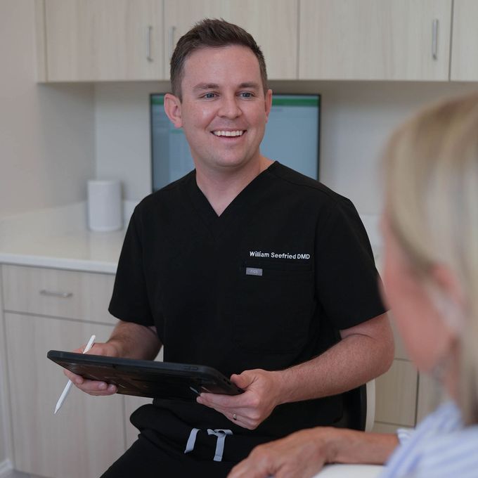 A man in a black scrub is holding a tablet and smiling