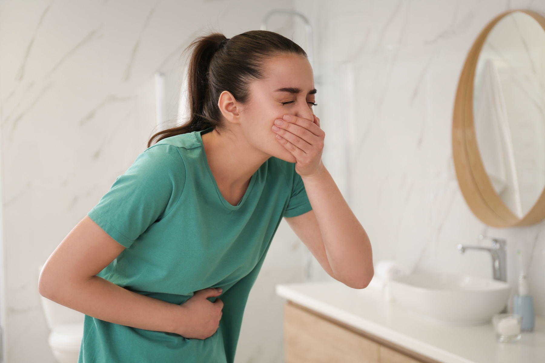 Person in teal shirt experiencing nausea in bathroom, covering mouth with one hand and holding stomach with other hand, indicating digestive distress