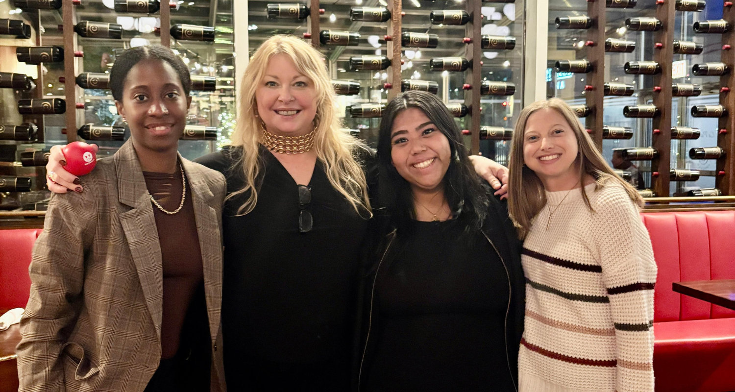 Four people smiling, posing together inside a restaurant. Wine bottles on the wall, red booth.