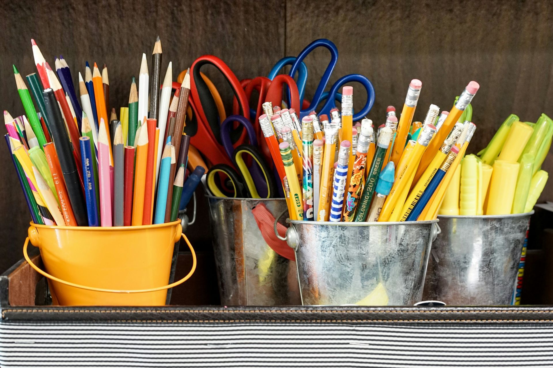 An assortment of educational supplies separated into 4 small tin buckets by type, including colored pencils, scissors, pencils, and highlighters.