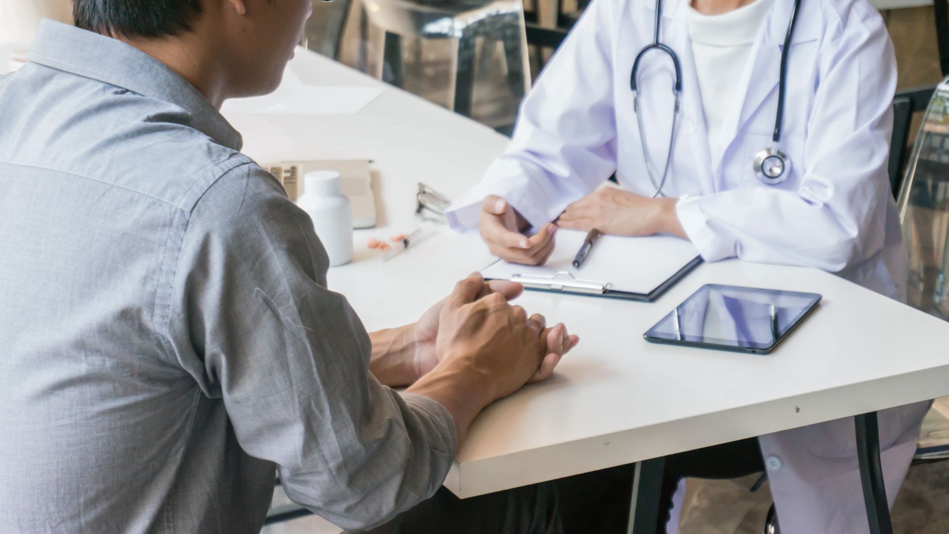 Doctor and patient at a desk, doctor in white coat writing on clipboard, patient facing doctor.