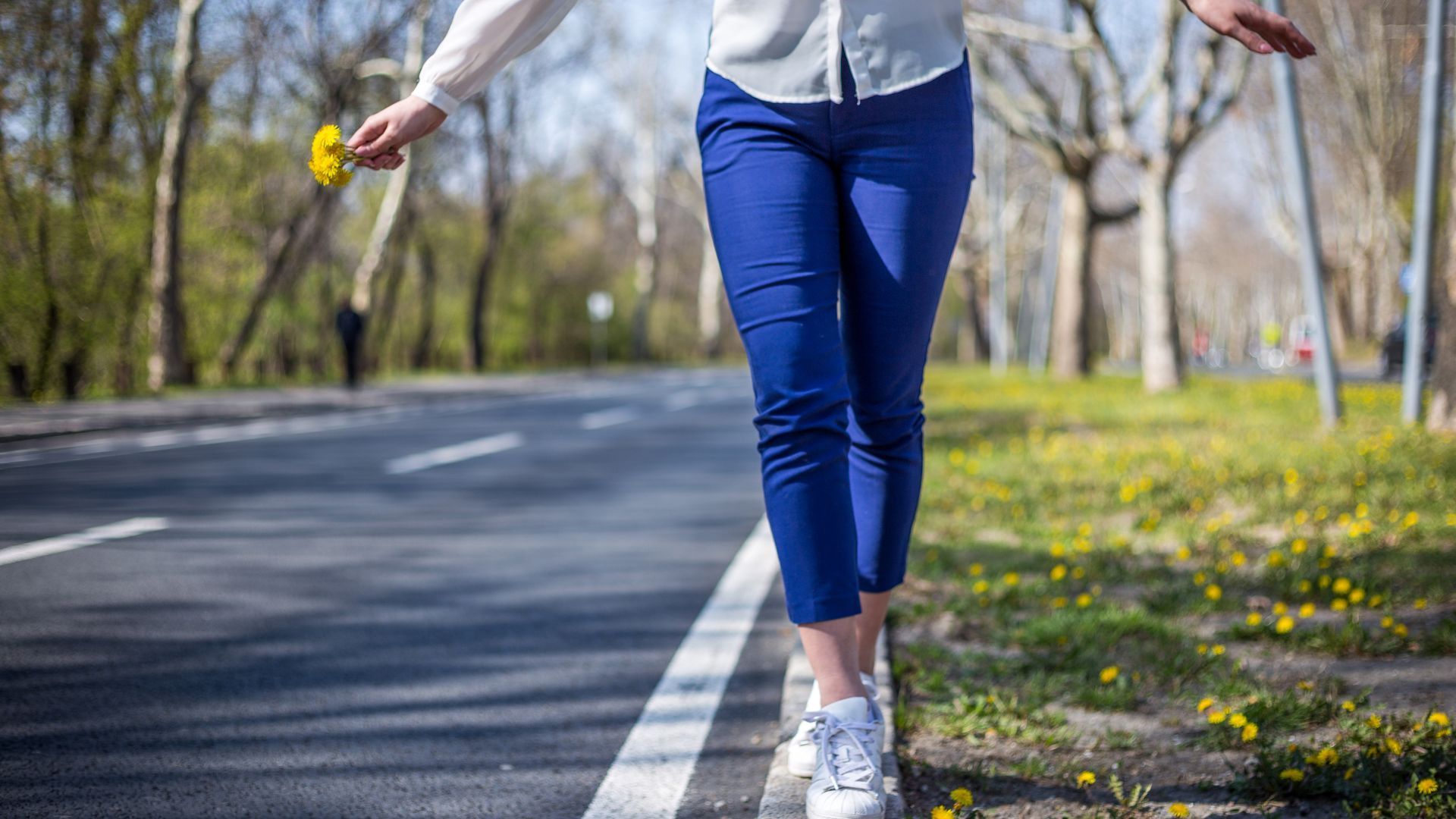 Woman balances on road curb, holding a yellow flower, wearing blue pants and white shoes, with trees in the background.