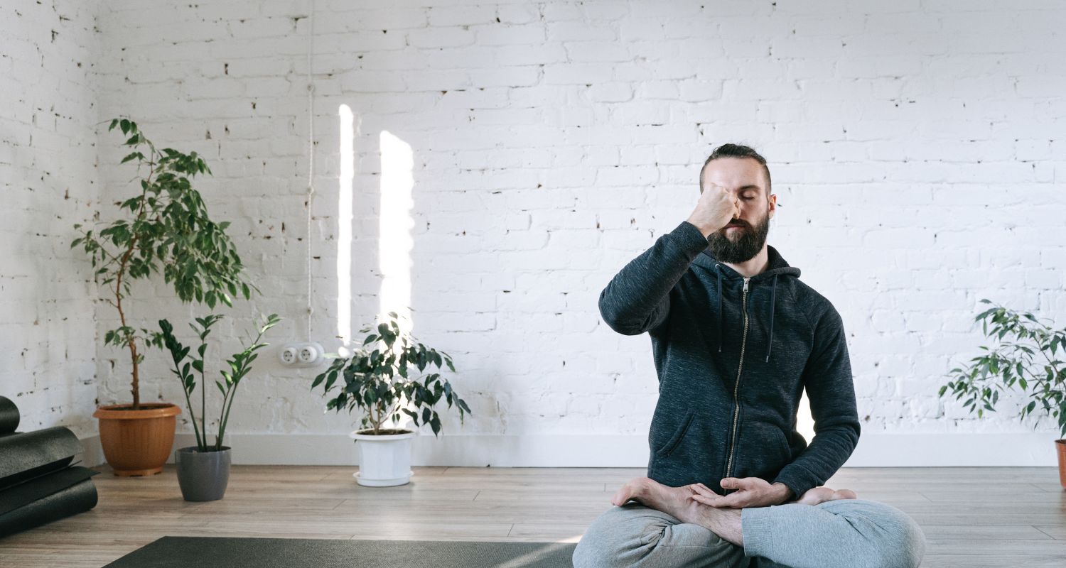 Man in yoga pose, practicing breathing technique indoors, white-walled room, plants, focused expression.