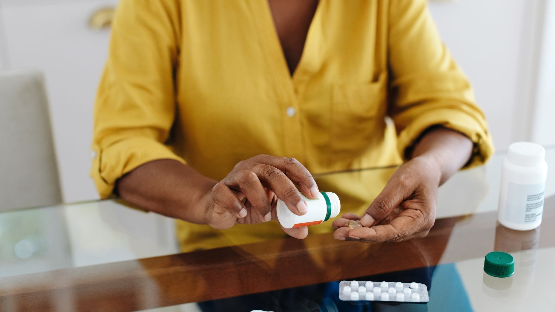 Person pouring pills from a white bottle into their hand at a table. Another pill bottle and blister pack are also visible.
