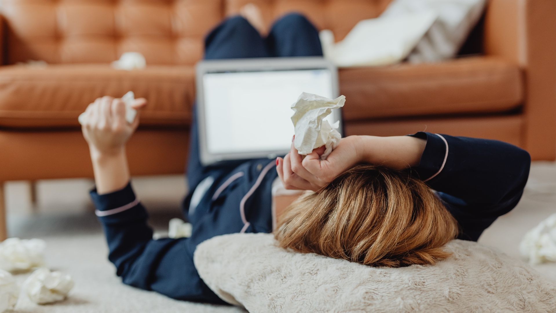 A person in dark pajamas lies on the floor with a laptop, surrounded by crumpled tissues, looking distressed.