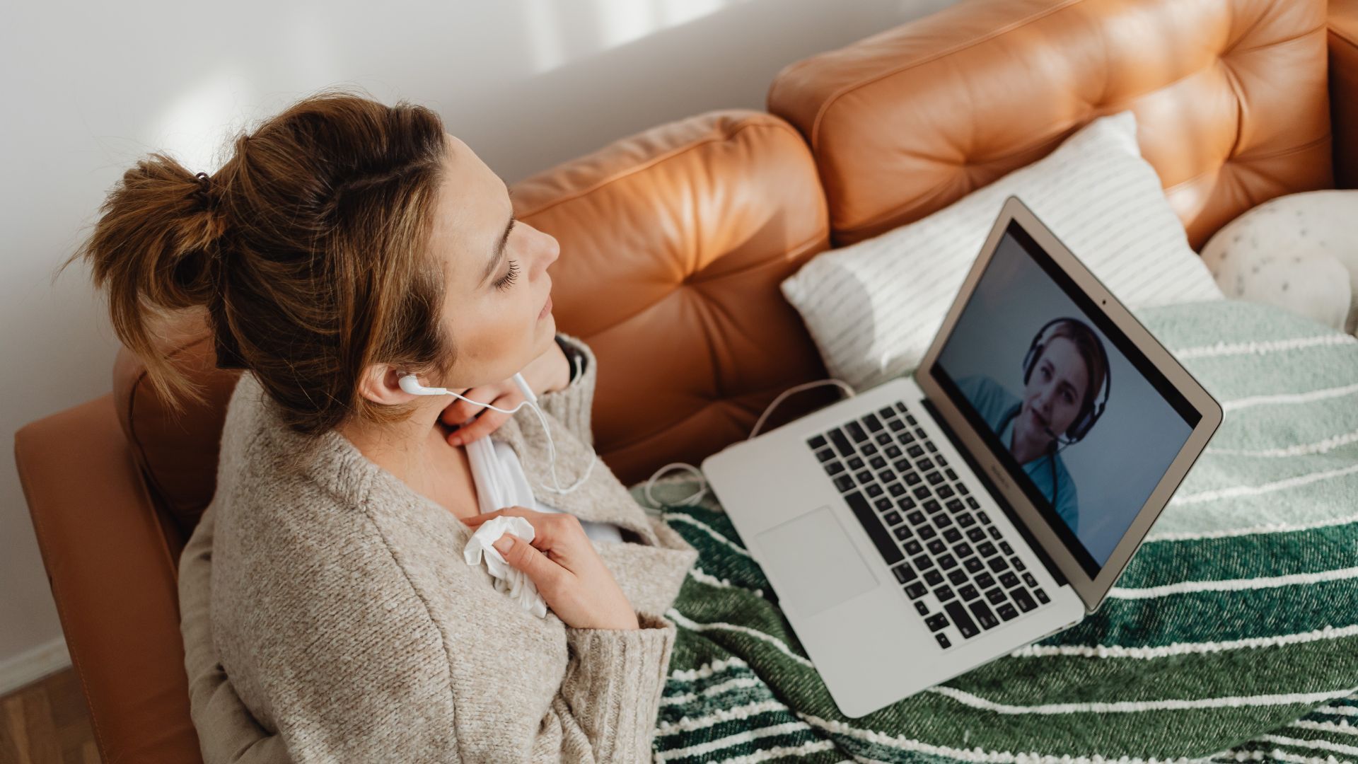 Woman on couch video chatting on laptop, wearing earbuds.
