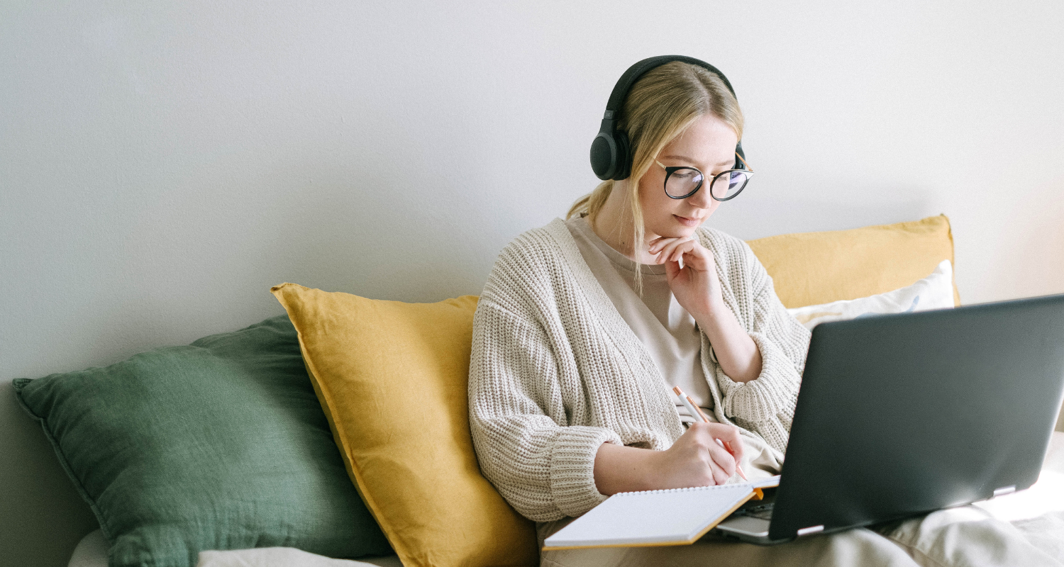 Woman wearing headphones and glasses, working on a laptop while taking notes on a couch with pillows.