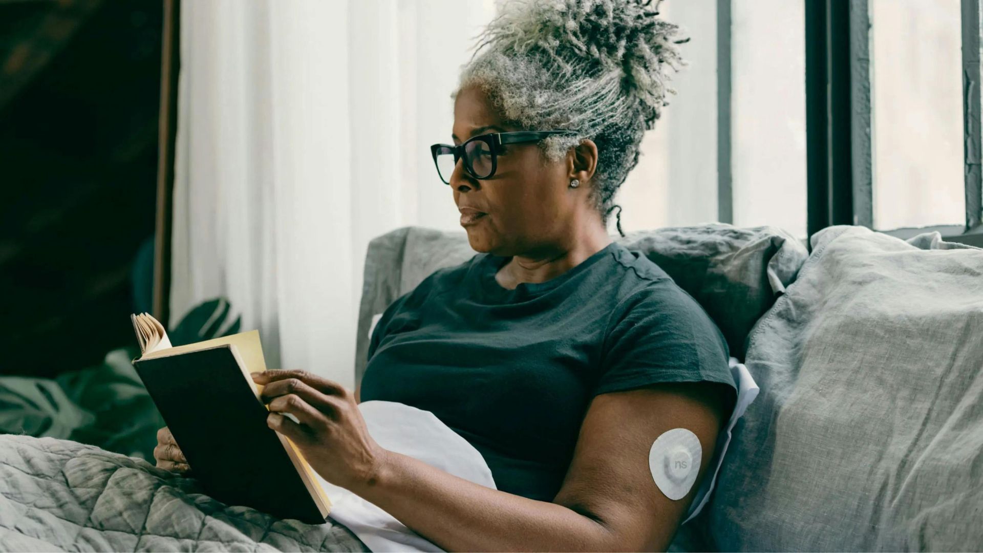 Woman reading a book on a couch, wearing glasses. An insulin pump is attached to her arm.