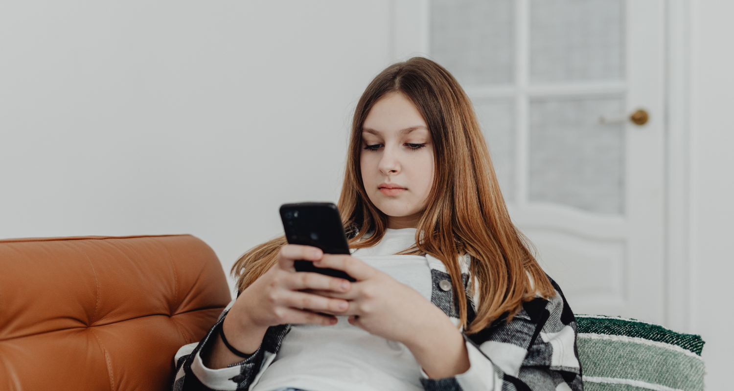 Girl with long hair, looking at a smartphone, sitting on a sofa in a room with a white door.