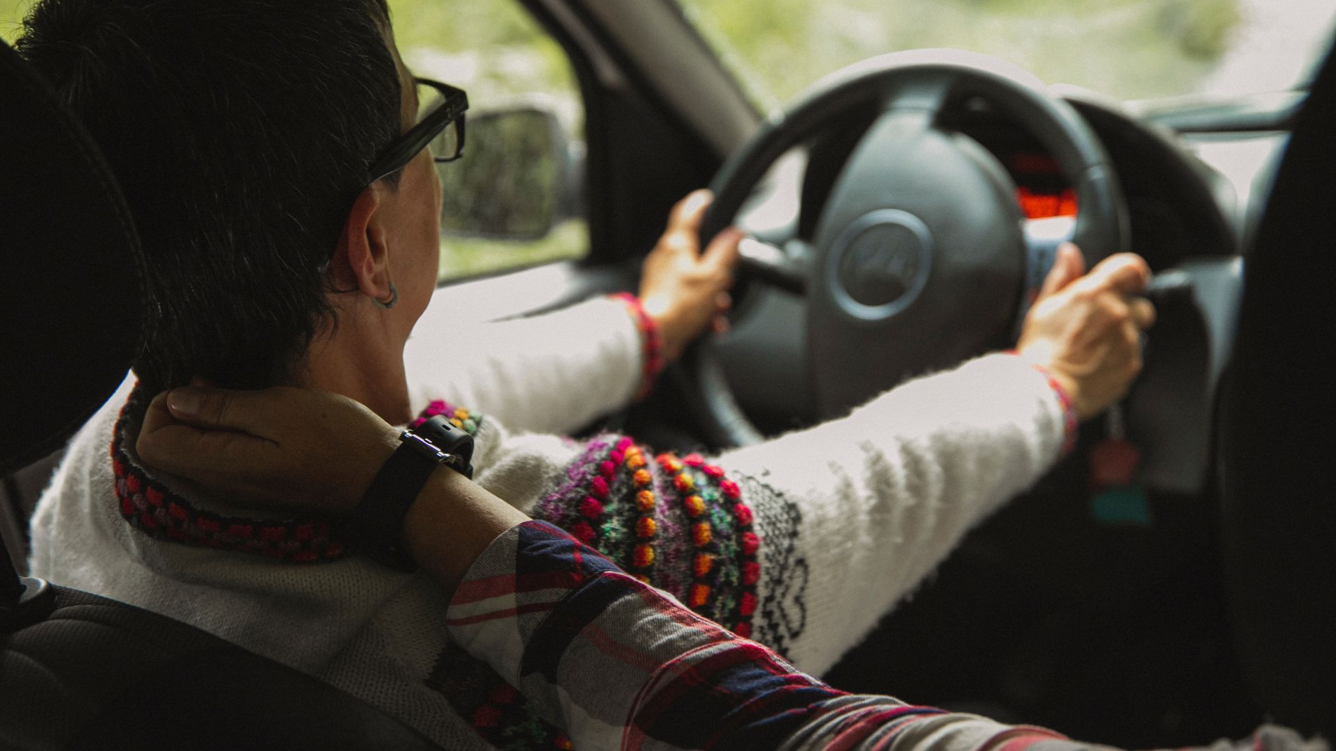Person driving, hands on steering wheel, another person's arm wrapped around their neck.