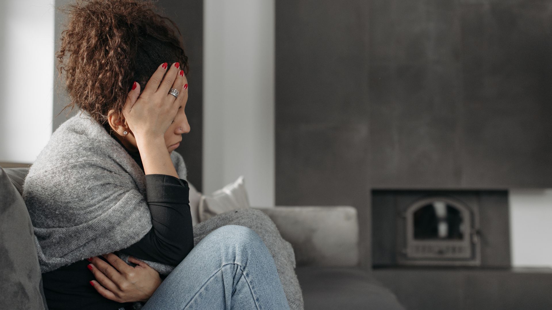 Woman, sad, sitting on couch, hand on head, wrapped in blanket, next to fireplace.
