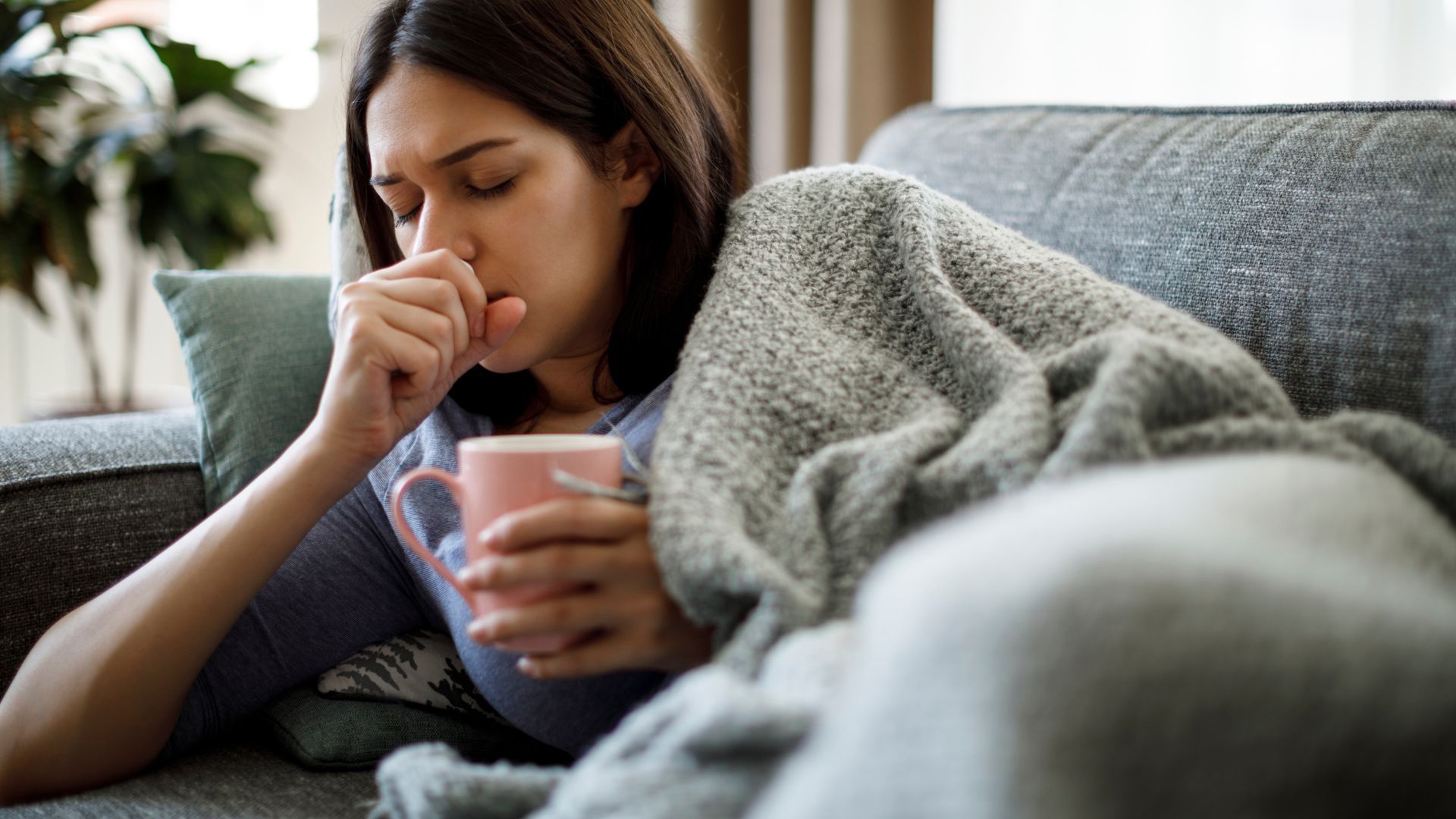 Woman on a couch, coughing and holding a mug, wrapped in a blanket.
