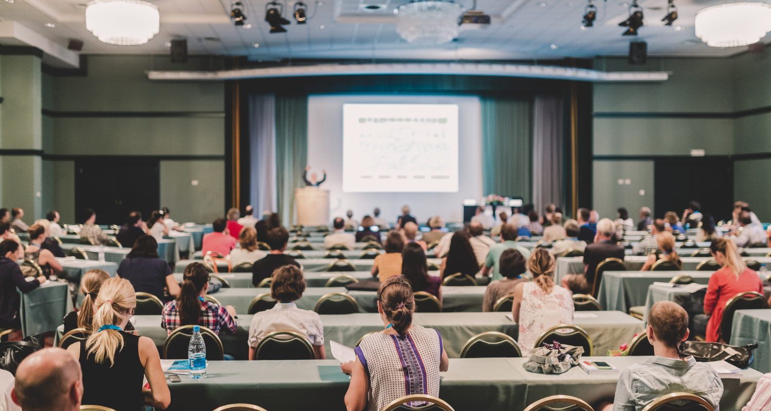 Large conference audience facing a projected presentation in a hotel ballroom conference hall.