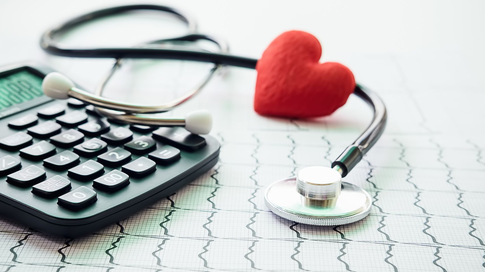 A calculator and stethoscope rest on a background of an EKG graph, with a small red heart figure placed near the tubes.