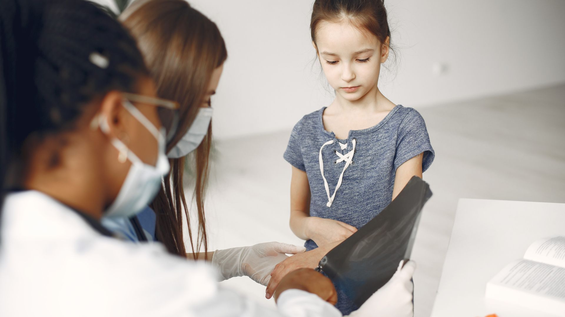 A doctor examines a child's arm in a medical setting, with the parent present, all wearing masks.