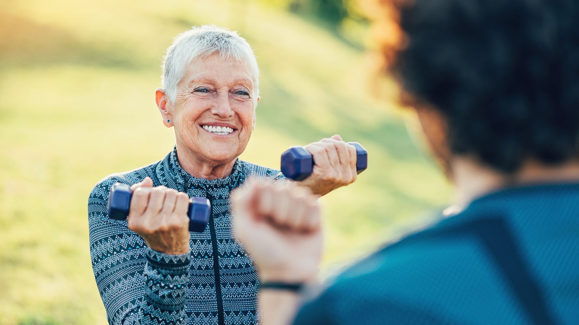 Woman smiles while lifting dumbbells with a person, outdoors.