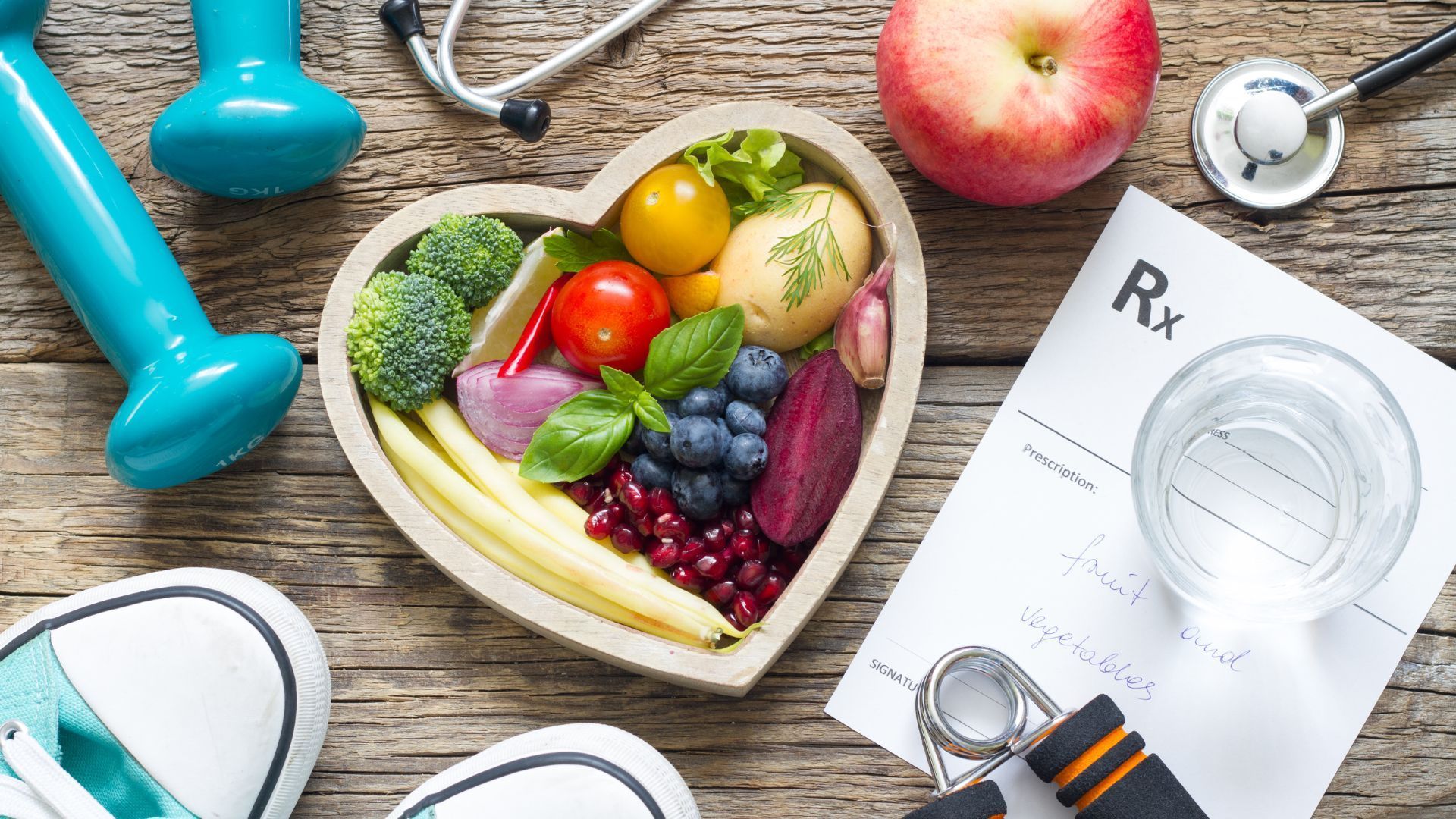 Heart-shaped bowl of vegetables, fruit, exercise equipment, prescription, and stethoscope on wood surface.