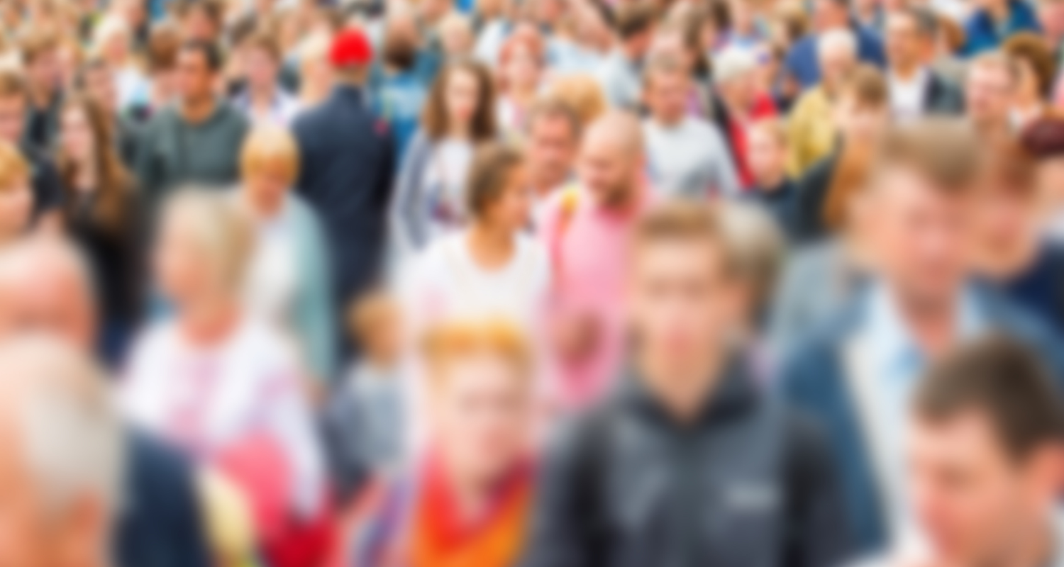 Blurred crowd of people walking outdoors on a busy street, many wearing colorful clothing.