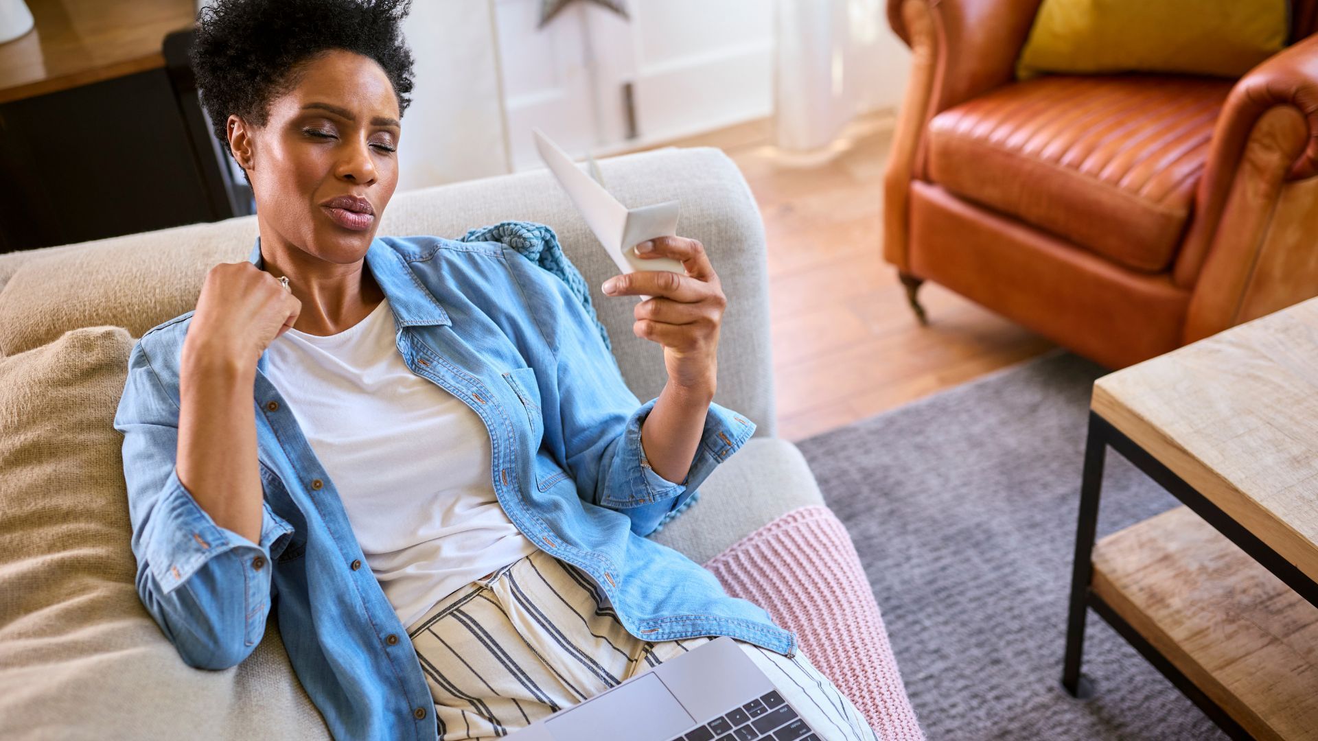 Woman on sofa using a laptop, holding paper, looking warm.