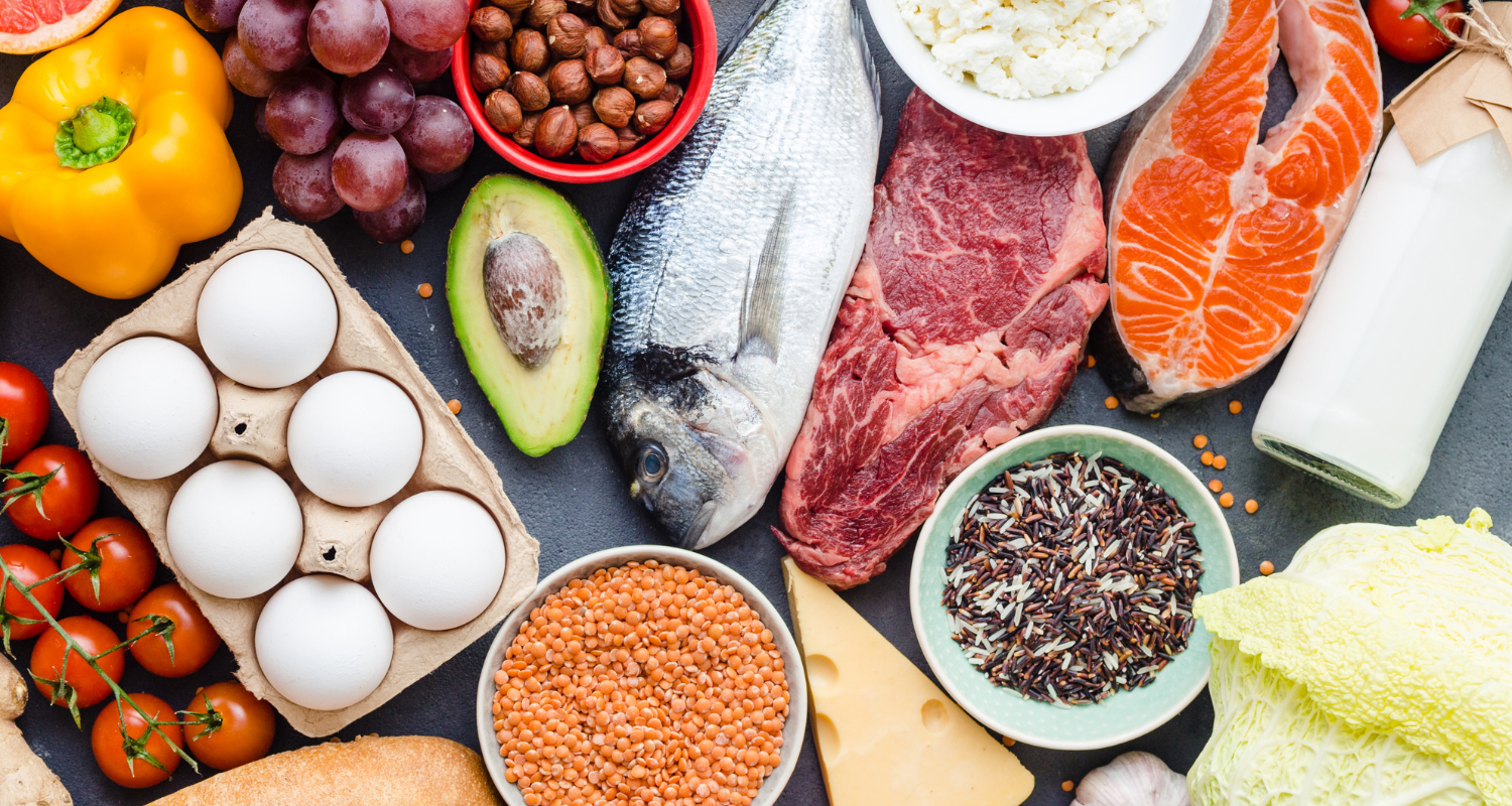 Assorted fresh foods including eggs, fish, meat, vegetables, fruit, grains, and dairy on a dark background