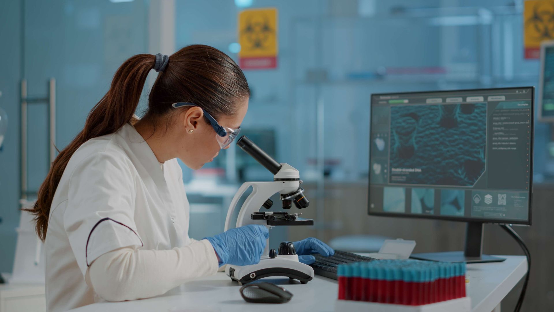A scientist in lab coat and gloves looks into a microscope, working in a laboratory with a computer.