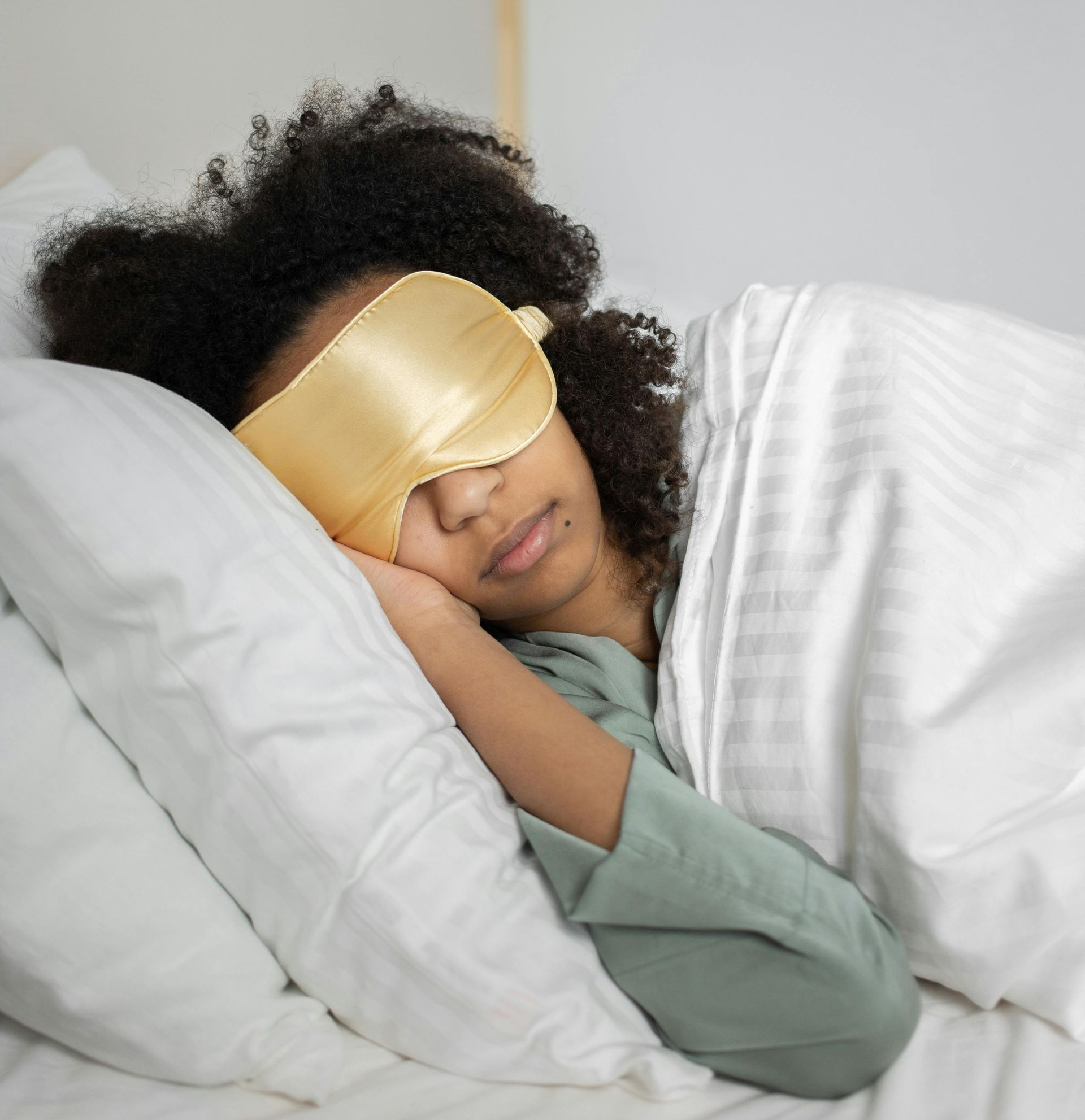 A person with dark curly hair sleeps in a bed wearing a gold satin eye mask, resting their head on a white pillow.