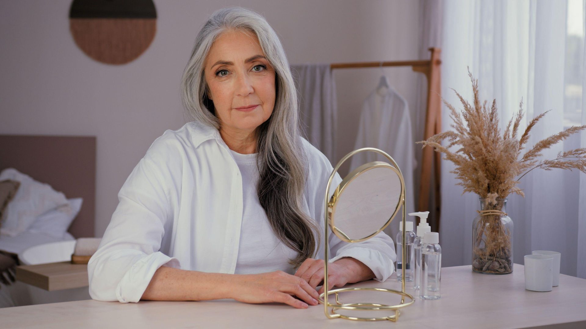Woman in a white blazer seated at a vanity, holding a makeup mirror in a softly lit room.