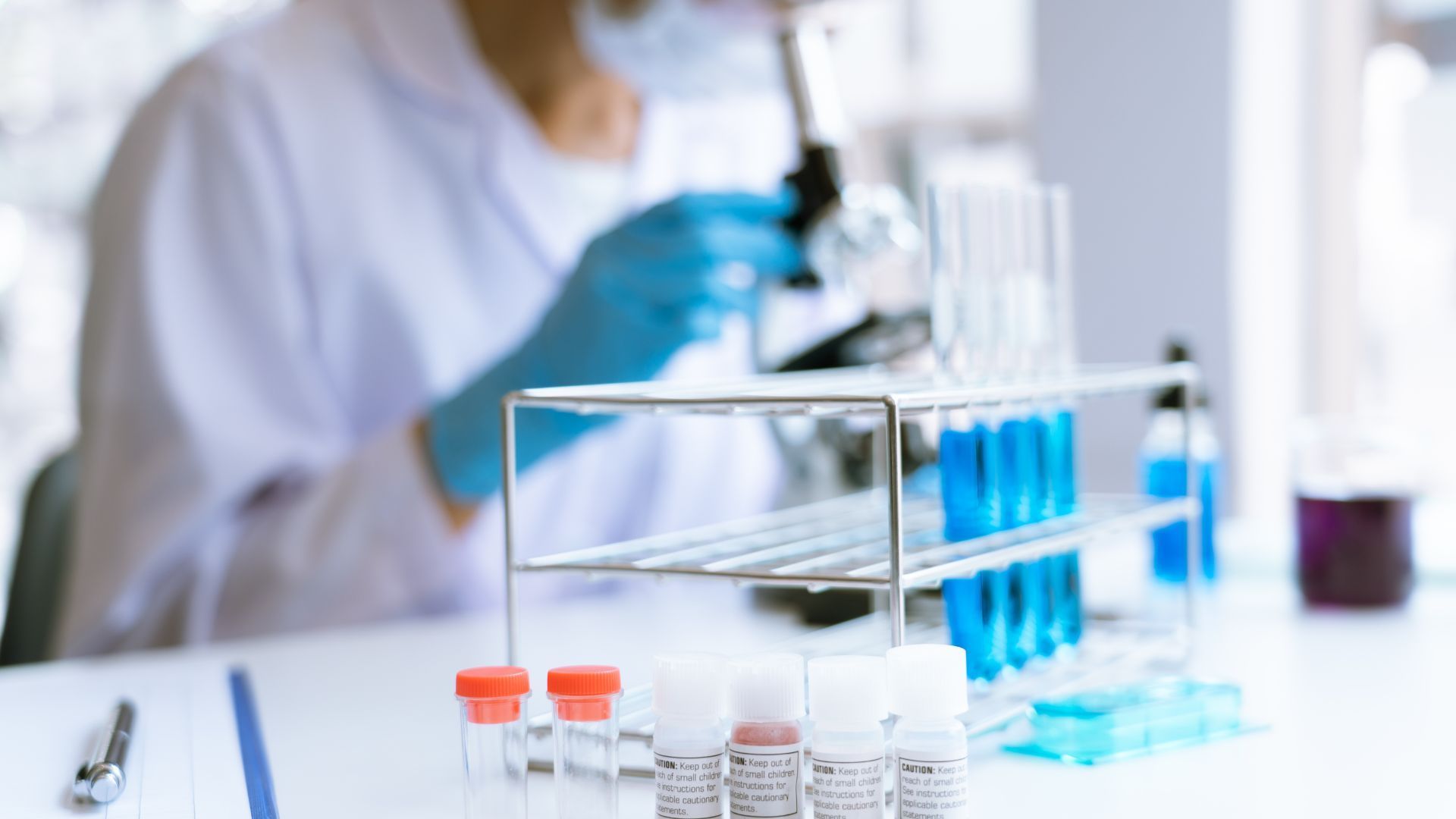 Scientist in lab coat and gloves looking into microscope; test tubes with blue liquid on rack.