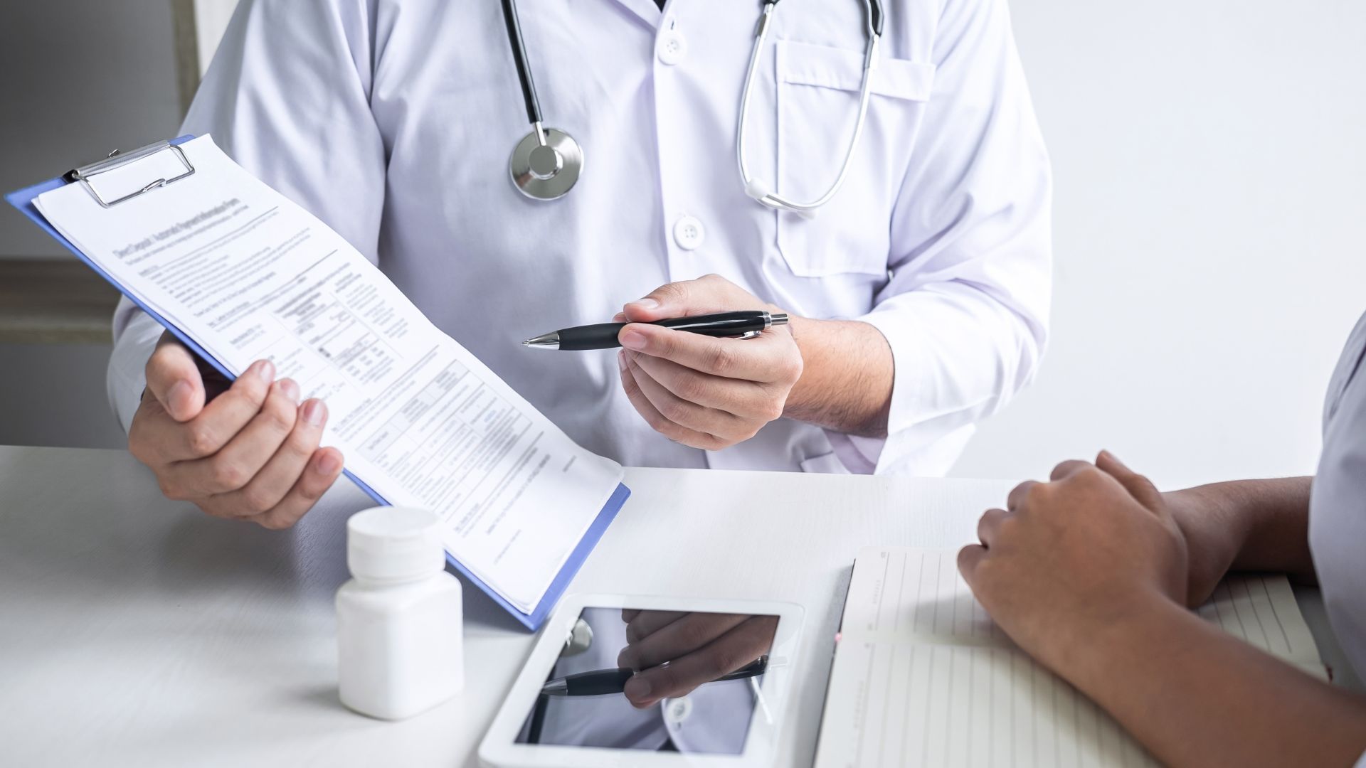 Doctor in white coat reviewing medical paperwork with a patient, pill bottle and tablet on the desk.