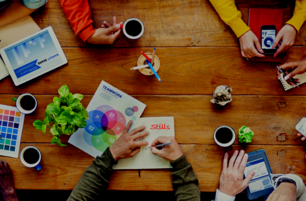 An overhead view of a wooden table with multiple people's hands visible working on documents, with coffee cups, a plant, and creative materials spread across the surface, representing collaborative resource development