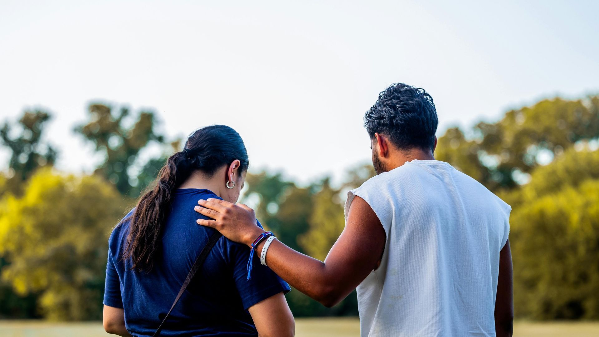 Man comforts a person by placing a hand on their shoulder outdoors in a park. Trees and sky in background.