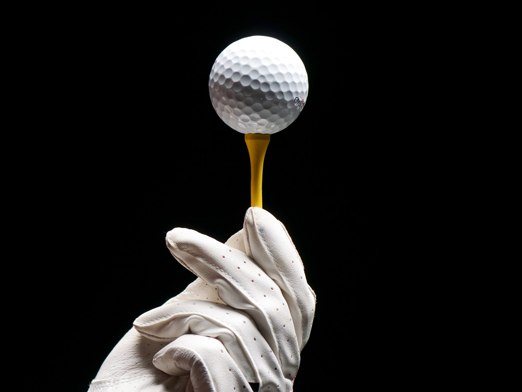 A hand in a white glove is holding a golf ball on a tee.