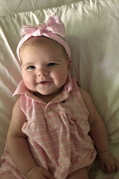 A baby girl wearing a pink dress and a pink headband is laying on a bed.