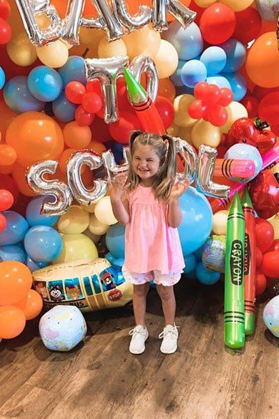 a little girl in a pink shirt smiling in front of a wall of balloons.