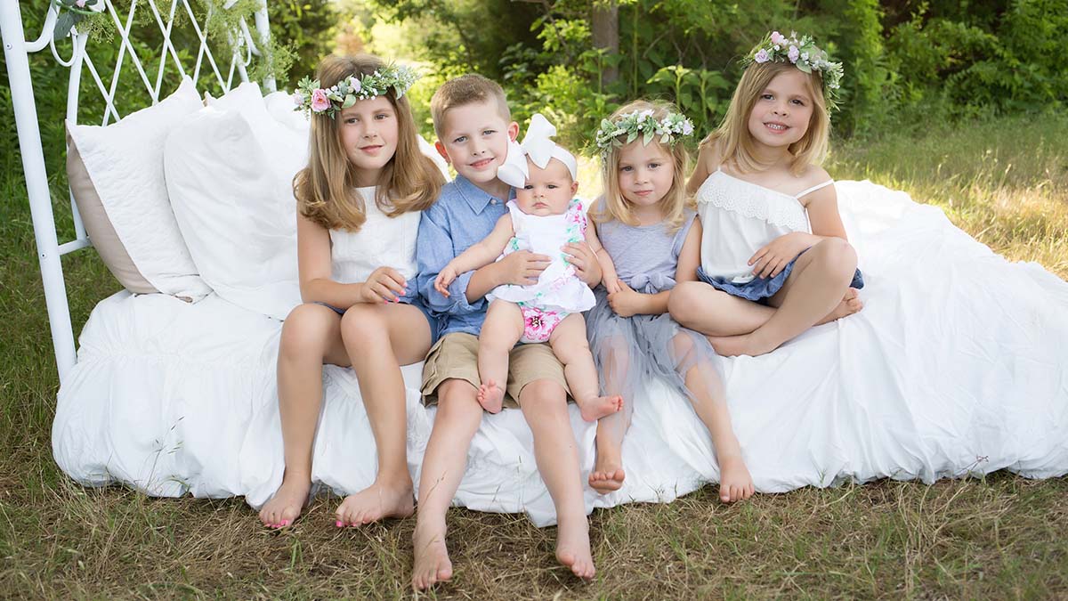 five young kids sitting on a white bed outside smiling for the camera