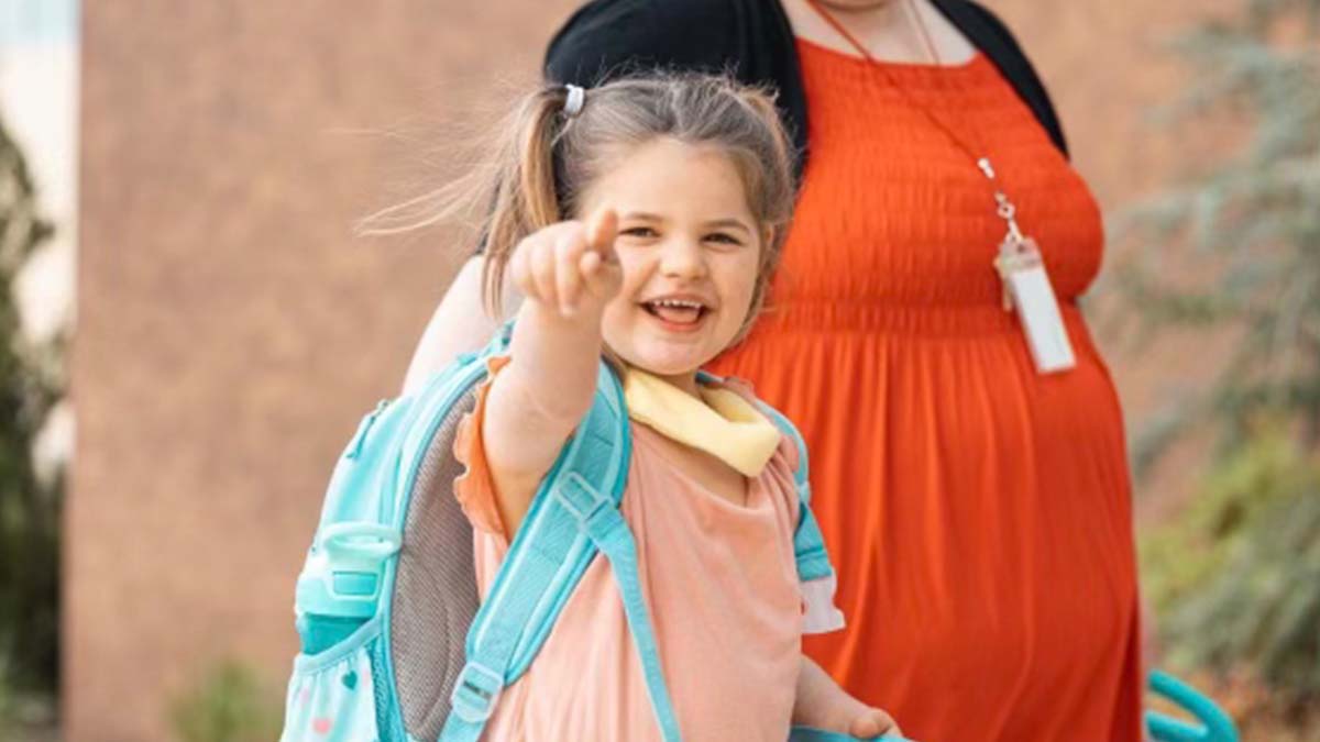 A little girl with a backpack is pointing at the camera while standing next to a woman.