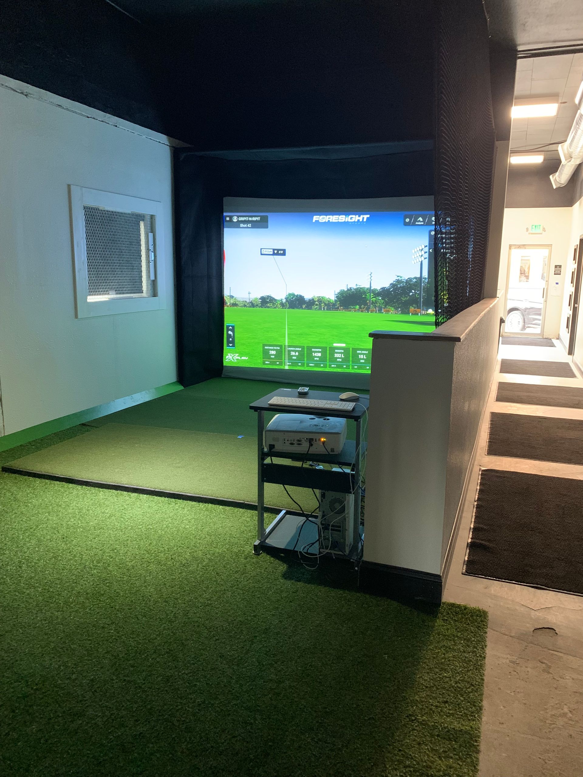 Golf simulator room, green turf, screen displaying course, equipment cart, white walls.