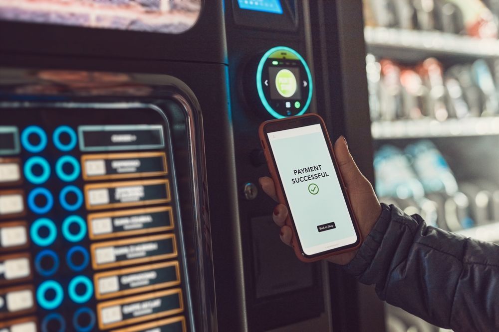 A person using a smartphone for contactless payment at a vending machine. Phone displays 