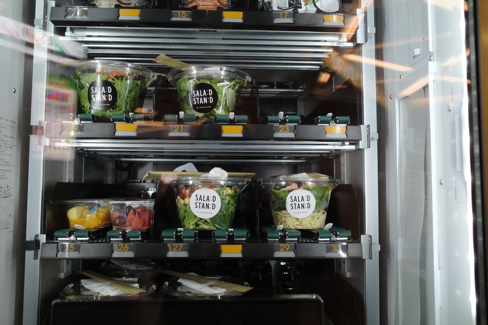 Vending machine stocked with pre-packaged salads, visible through glass. Green, white, and clear containers on shelves.