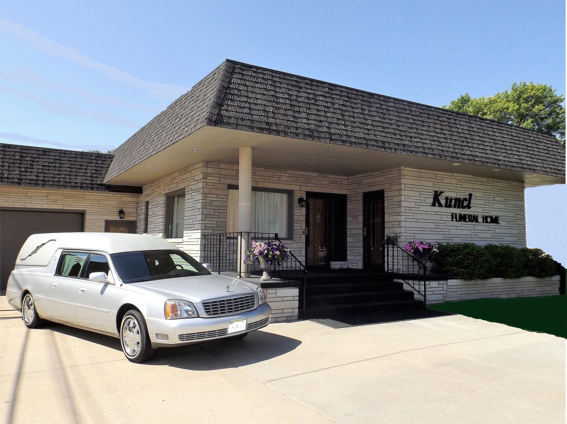 A silver hearse parked in front of a light-colored brick funeral home under a clear blue sky.