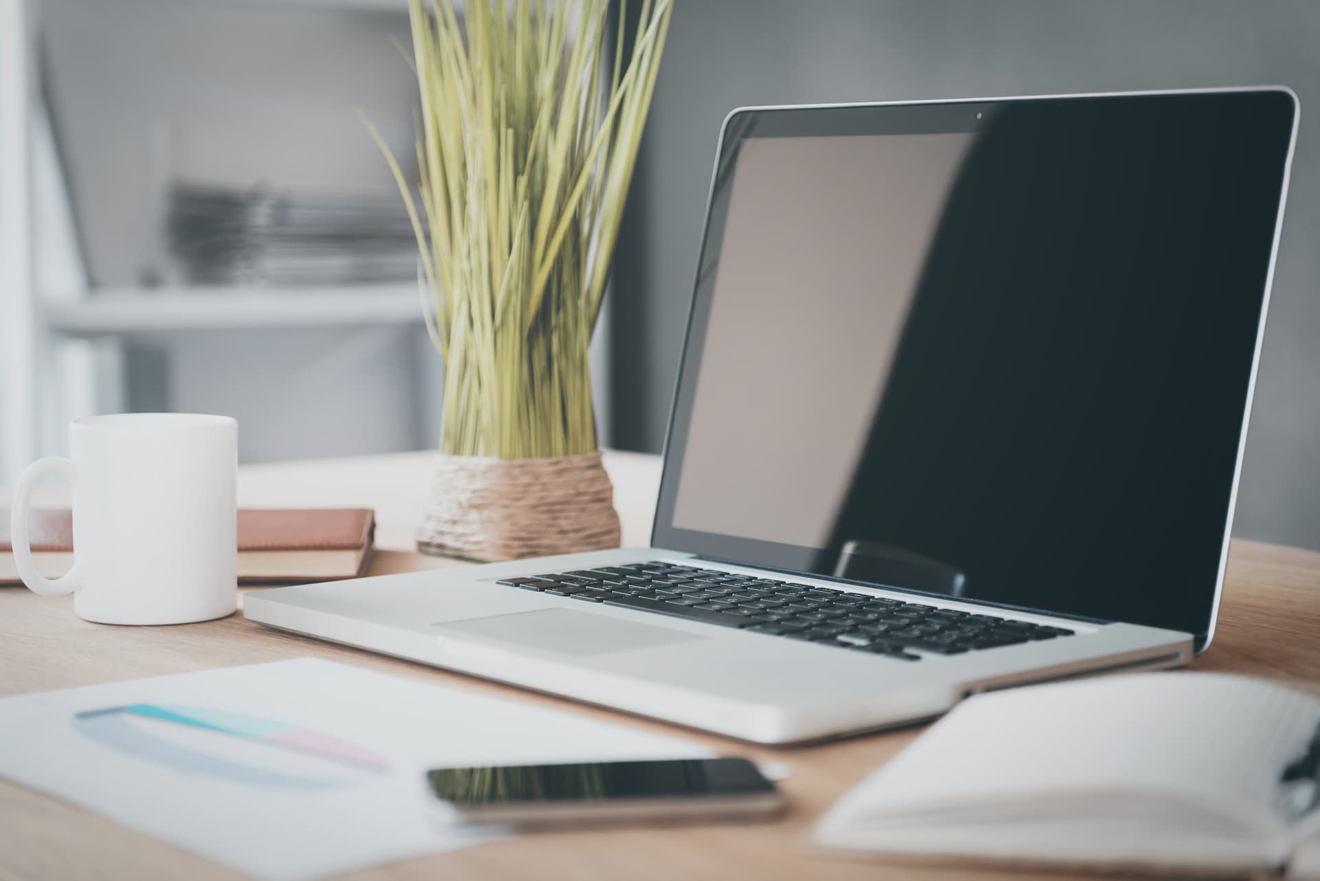 A laptop, white mug, smartphone, and potted grass on a wooden desk, with a document and a closed notebook nearby.