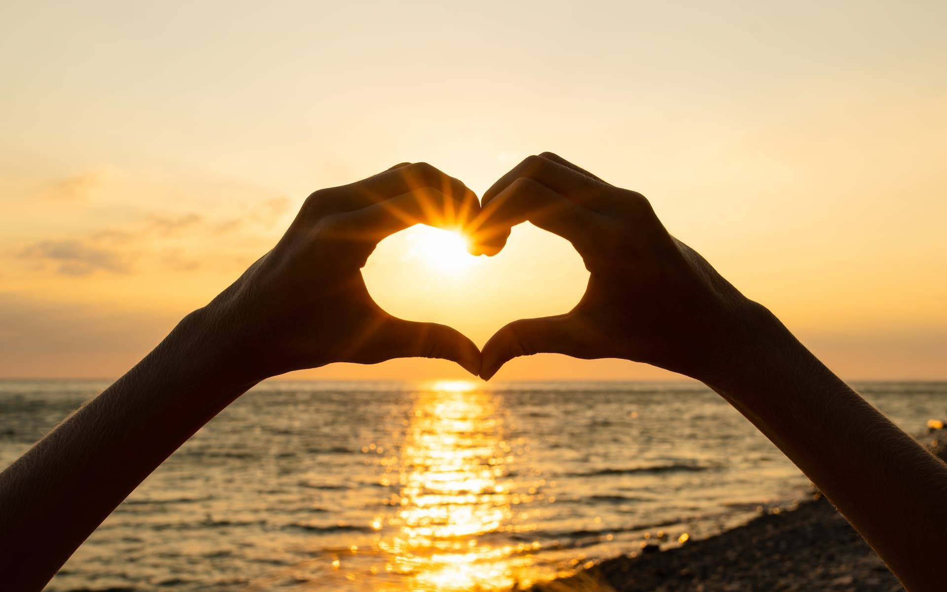 Hands forming a heart shape against a golden sunset over the ocean.