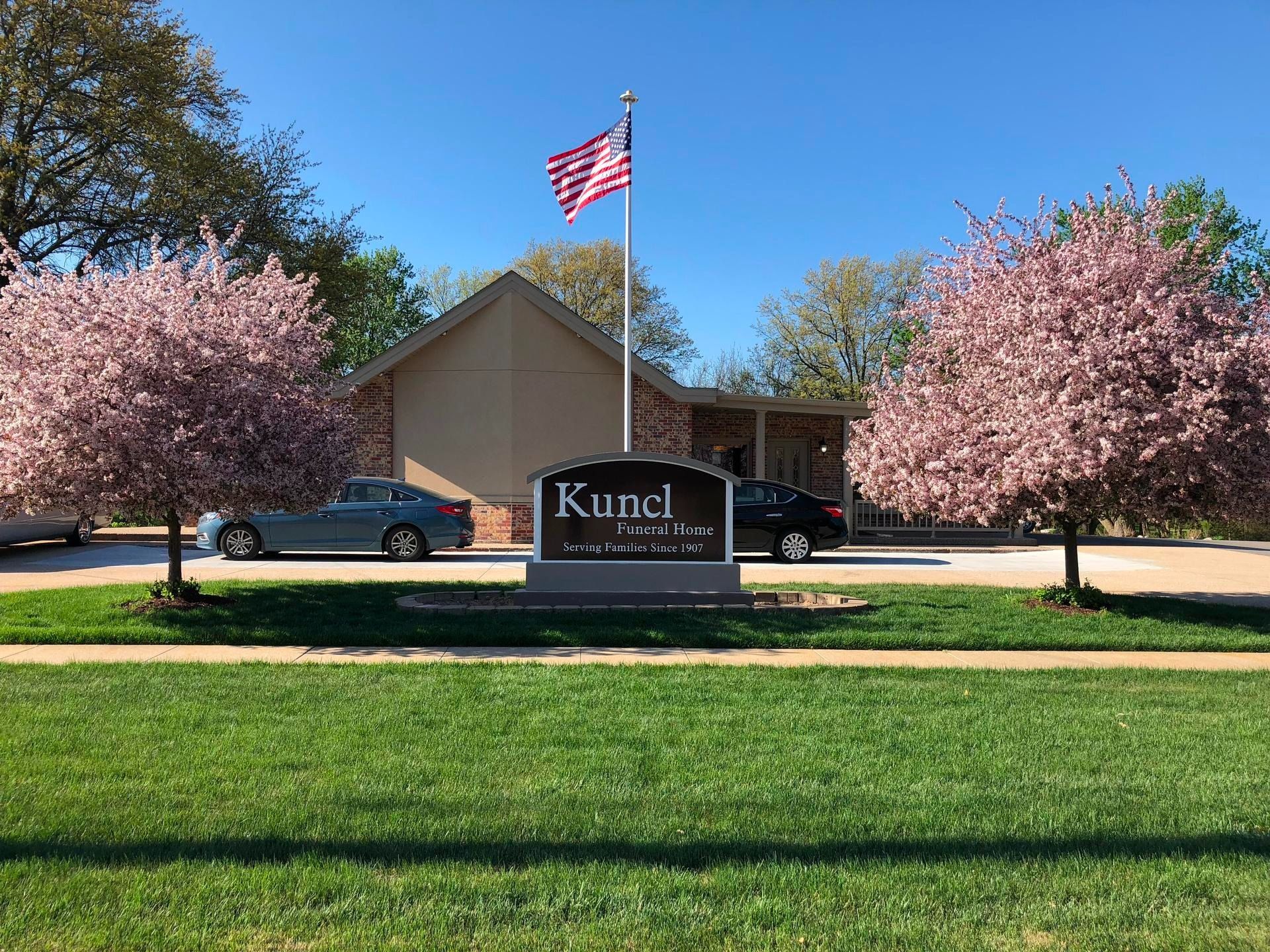 Kuncl Funeral Home sign in front of a building with two blooming pink trees and an American flag under a blue sky.
