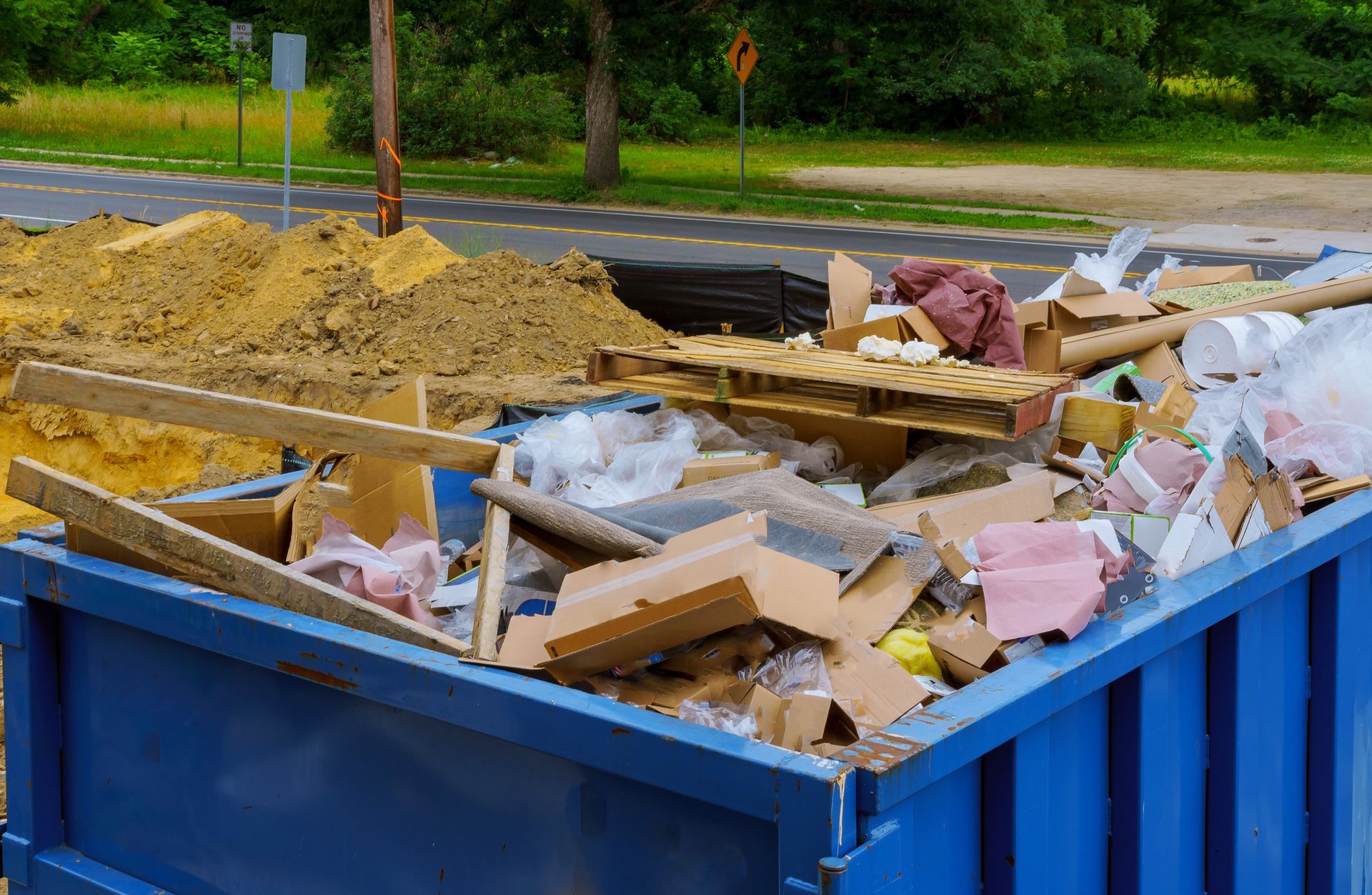 Blue dumpster overflowing with construction debris, dirt pile, and street in background.