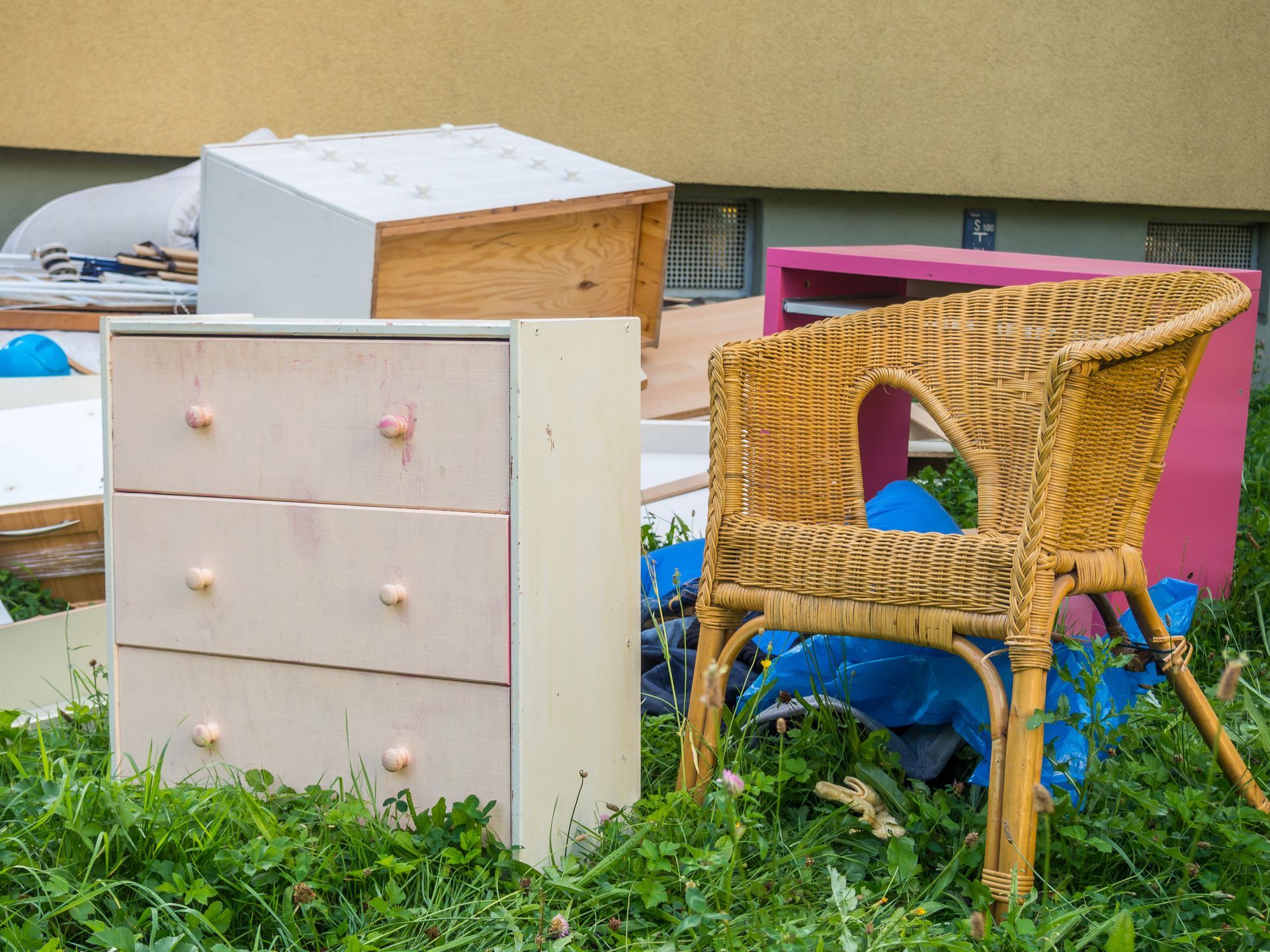 Discarded furniture on grass, including a dresser, wicker chair, and other pieces in various colors.