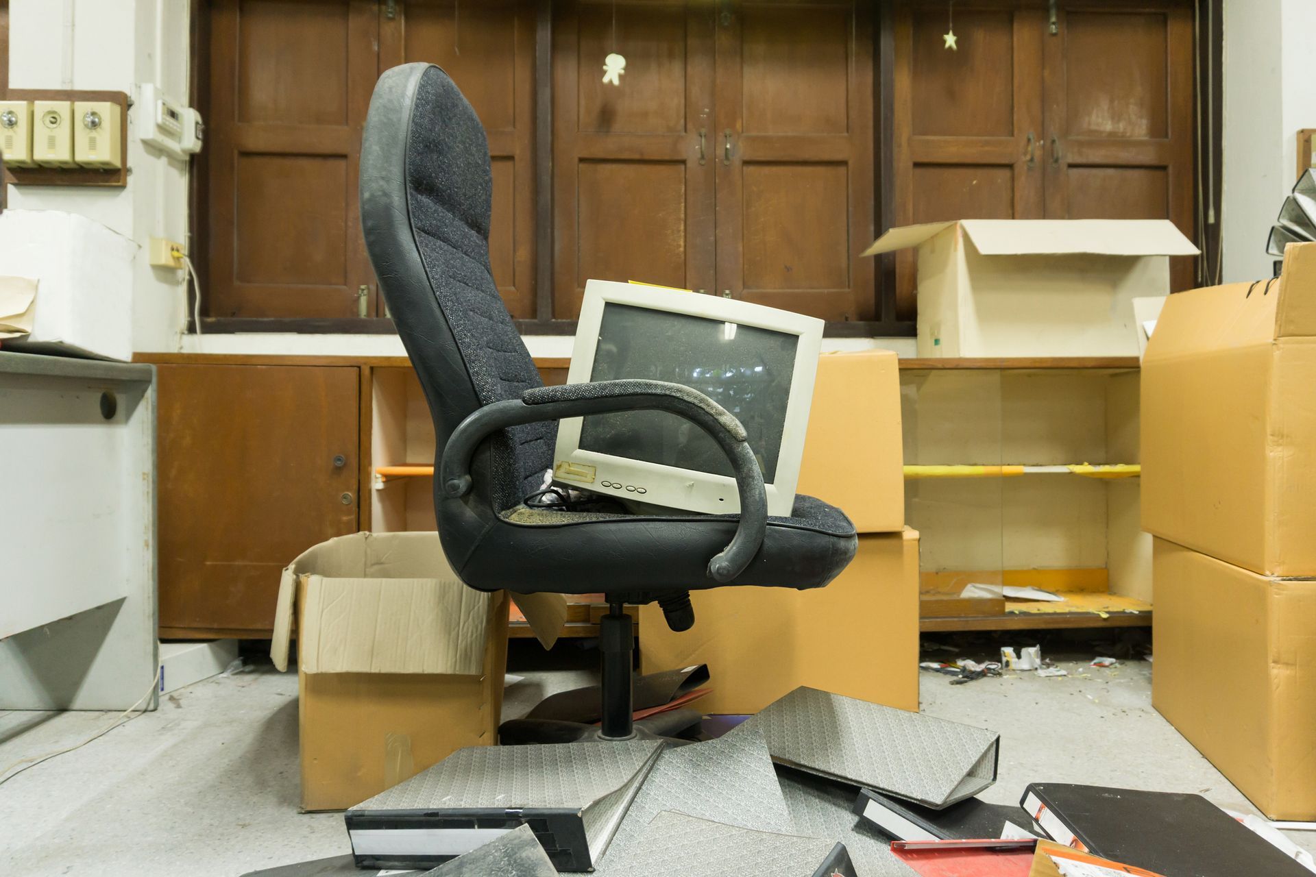 Office desk and chair in disarray, with an old computer monitor atop the chair. Boxes and books are scattered.