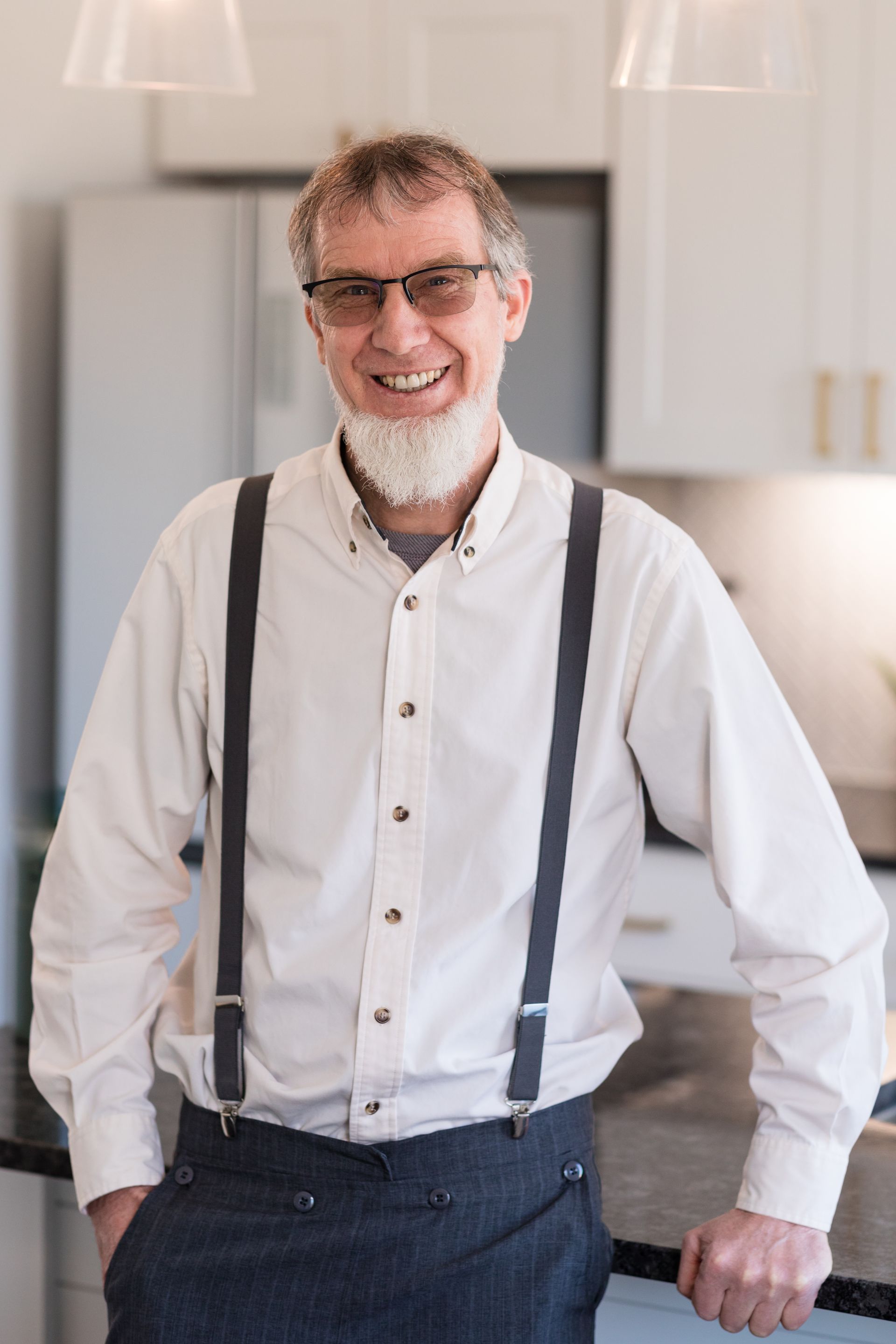 A man with a beard and suspenders is standing in a kitchen.